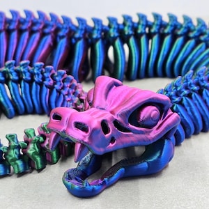 May include: A 3D-printed articulated snake toy in iridescent blue, purple, and green. The snake's head is open, showing teeth. The body is segmented for flexible movement. The toy is designed to resemble a dragon or serpent.