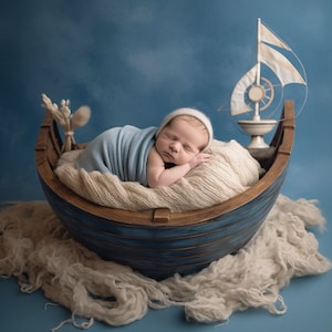May include: A newborn baby sleeping in a blue and brown wooden boat-shaped prop, wrapped in a blue blanket and a white knit hat. The boat is surrounded by white fluffy fabric, and a small white sailboat is in the background.