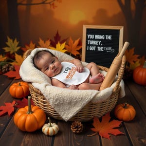 May include: A baby wearing a white bib with the text "I toy I am thankful for you" in orange and black lettering, lying in a wicker basket with a rolling pin and pumpkins. The background is a fall-themed backdrop with orange, red, and yellow leaves.