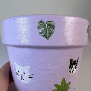 以下が含まれることがあります： 手描き模様のラベンダー色のセラミックプランター。プランターには、緑色のモンステラの葉、ピンクの鼻をした白い猫、白黒の猫、白いドットのある緑色の植物が描かれています。