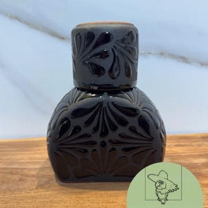 May include: A black ceramic vessel with a wide, rounded base and a cylindrical top, featuring a glossy, raised floral pattern carved into the matte surface. The piece has a distinct terracotta-colored rim visible at the very top of the object.