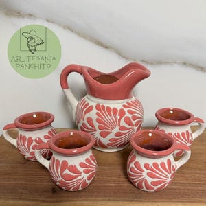 May include: A set of five hand-painted ceramic pottery pieces in a white and pink color scheme. The set includes a pitcher and four small cups. The cups and pitcher feature a floral design with pink accents.
