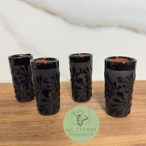 May include: Four black ceramic shot glasses with intricate floral designs. The glasses are arranged in a row on a wooden surface.