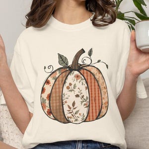 May include: A cream-colored short-sleeve t-shirt featuring a large central graphic of a pumpkin. The pumpkin is divided into vertical segments with alternating patterns, including orange plaid, solid brown, and floral prints in shades of orange and green.