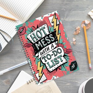 May include: A spiral-bound notebook with a colorful cover featuring the text "HOT MESS WITH A TO-DO LIST." The design includes red, teal, and yellow accents, with illustrations of lips and lightning bolts. The notebook is on a wooden surface.