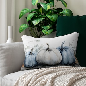 May include: A rectangular throw pillow featuring a watercolor illustration of three pumpkins in shades of muted blue and white. The design includes delicate vine and leaf outlines in the background, set against a soft, off-white fabric base.