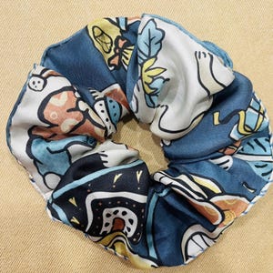 May include: A patterned scrunchie featuring a repeating design of cartoon-like figures and floral elements in shades of blue, white, yellow, and orange. The fabric is gathered and sewn to create a circular shape, perfect for securing hair.