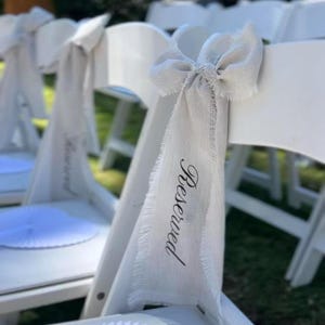 Op de afbeelding: Witte klapstoelen versierd met lichtbeige stoffen strikken en "Gereserveerd" banners. De banners hebben een gerafelde rand en zijn gedrukt in een cursief lettertype. De stoelen staan buiten opgesteld, klaar voor een evenement.