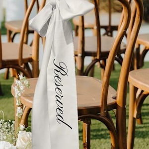 Op de afbeelding: Houten stoelen met gebogen rugleuningen, buiten opgesteld. Een witte satijnen sjerp met het woord "Reserved" in zwart script is aan de achterkant van een stoel vastgebonden. De scène suggereert een bruiloft of een speciale gebeurtenis.