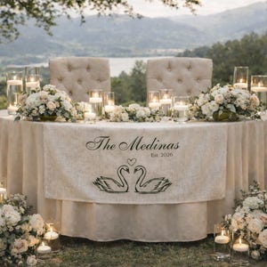 Könnte beinhalten: Ein Hochzeitsempfangstisch mit cremefarbener Tischdecke und zwei getufteten Stühlen. Der Tisch ist mit Blumenarrangements, Kerzen und einem Banner mit der Aufschrift "The Medinas Est. 2026" mit Schwanenillustrationen dekoriert.