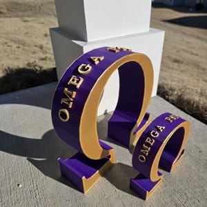 May include: Three-dimensional purple and gold Omega Psi Phi fraternity symbols. The symbols are in the shape of the Greek letter Omega. The text "OMEGA" is visible on the largest symbol. The symbols are on a concrete surface.