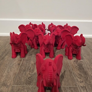 May include: A collection of red elephant figurines, each with detailed features like large ears, a trunk, and a decorative necklace. The elephants are arranged on a wooden surface, with one in the foreground and others in the background.