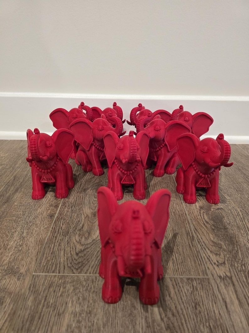 May include: A collection of red elephant figurines, each with detailed features like large ears, a trunk, and a decorative necklace. The elephants are arranged on a wooden surface, with one in the foreground and others in the background.