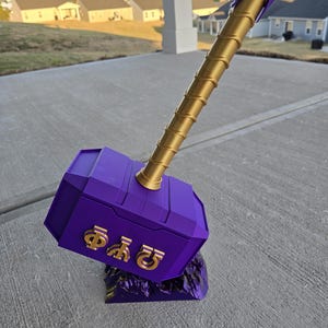 May include: A purple and gold decorative hammer with a textured gold handle and a purple head. The head features gold symbols. The hammer rests on a purple base, suggesting a fantasy or cosplay theme.