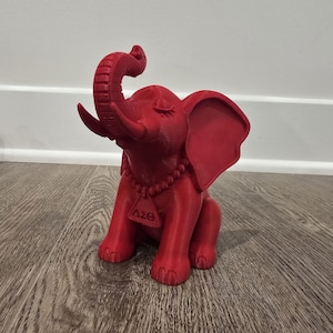 May include: A red elephant figurine with its trunk raised. The figurine has large ears, a beaded necklace, and a tag with Greek letters. The elephant is sitting on a wooden surface.