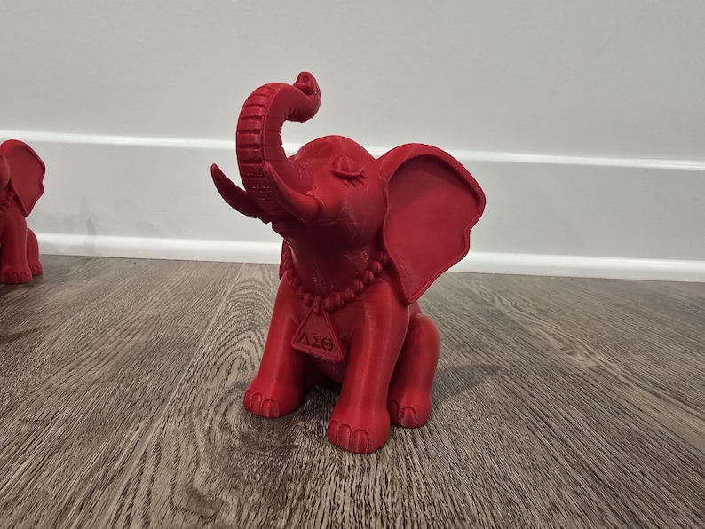 May include: A red elephant figurine with its trunk raised. The figurine has large ears, a beaded necklace, and a tag with Greek letters. The elephant is sitting on a wooden surface.