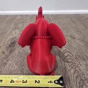 May include: A bright red elephant figurine, viewed from the back, with large ears and a detailed trunk. The elephant is sitting, and a measuring tape is in the foreground. The figurine is approximately 3 inches long.