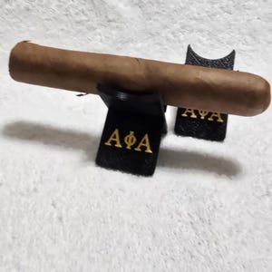 Peut inclure: Un cigare brun repose sur deux supports à cigares noirs. Les supports portent des lettres dorées qui se lisent "AΦA" et "AYA". Le cigare est de couleur brun clair. Le fond est une surface blanche et texturée.