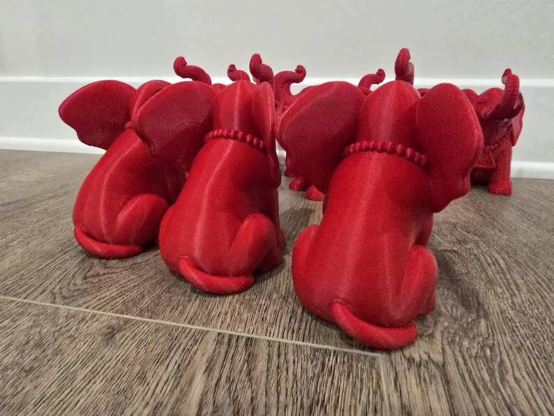 May include: A collection of red elephant figurines, each in a seated position with their trunks raised. The elephants have detailed features, including large ears and beaded necklaces. The figurines are arranged on a wooden surface.