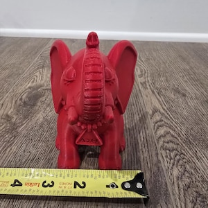 May include: A red elephant figurine with large ears, a raised trunk, and a decorative pendant. The figurine is approximately 3 inches tall, as measured by a yellow tape measure.