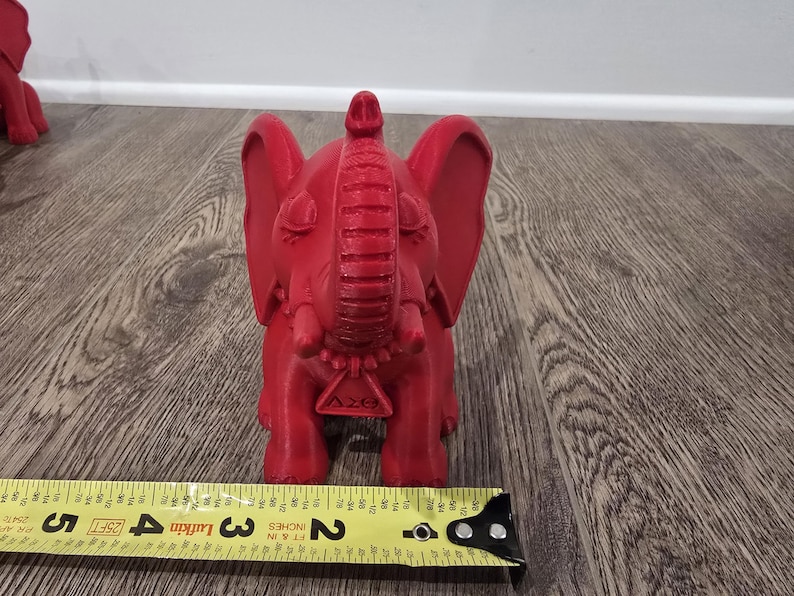 May include: A red elephant figurine with large ears, a raised trunk, and a decorative pendant. The figurine is approximately 3 inches tall, as measured by a yellow tape measure.