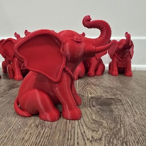 May include: A collection of red elephant figurines. The elephants are in a sitting position with their trunks raised. Each elephant has a necklace and a pendant. The figurines are displayed on a wooden surface.