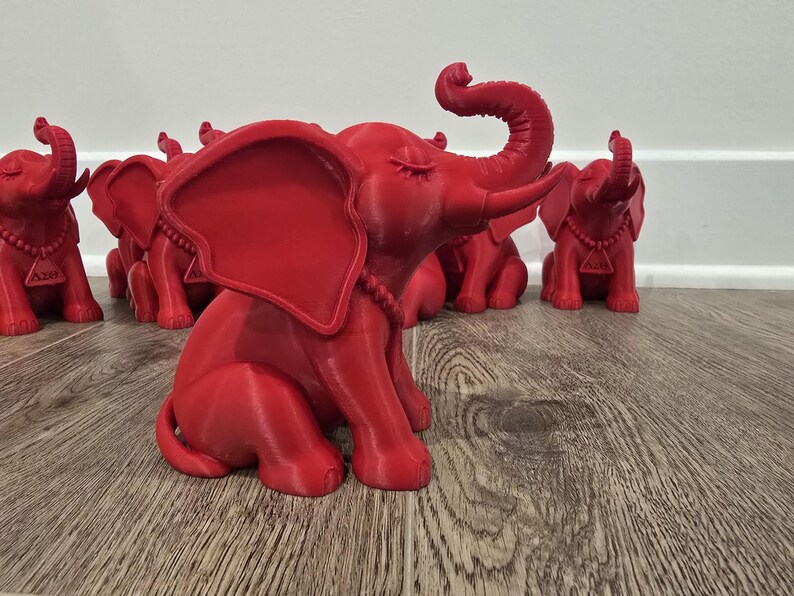 May include: A collection of red elephant figurines. The elephants are in a sitting position with their trunks raised. Each elephant has a necklace and a pendant. The figurines are displayed on a wooden surface.