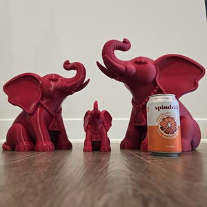 May include: Three matte magenta elephant figurines in varying sizes, each wearing a beaded necklace with a triangular pendant featuring the Greek letters Delta Sigma Theta. A 12-ounce Spindrift Blood Orange sparkling water can is placed for scale.