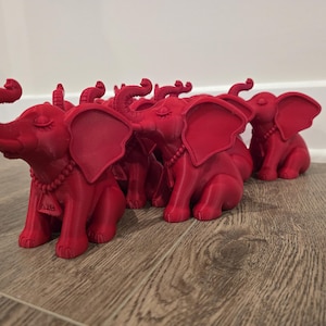 May include: A group of red elephant figurines with detailed features, including large ears, curled trunks, and beaded necklaces. The elephants are arranged on a wooden surface, with a white wall in the background. Each elephant has a small tag with the letters "A18".