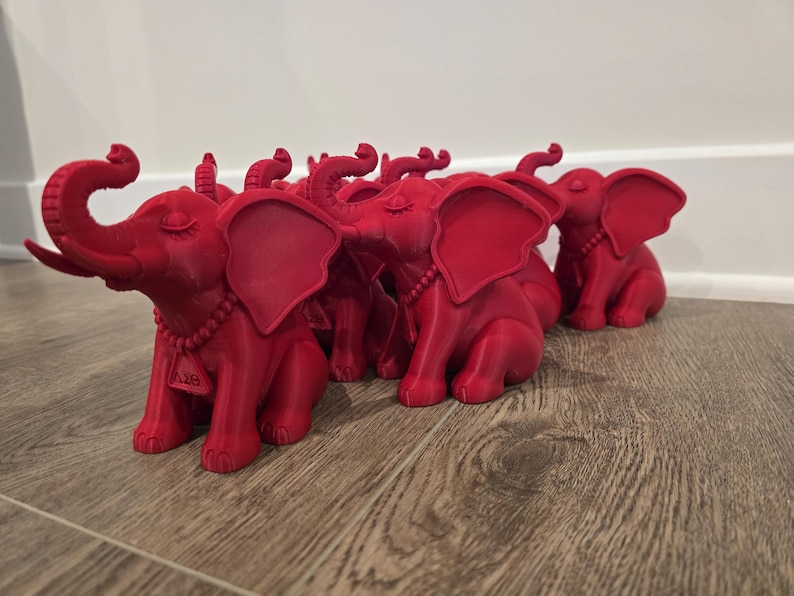 May include: A group of red elephant figurines with detailed features, including large ears, curled trunks, and beaded necklaces. The elephants are arranged on a wooden surface, with a white wall in the background. Each elephant has a small tag with the letters "A18".