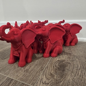 May include: A collection of red elephant figurines. Each figurine has large ears, a curled trunk, and a beaded necklace. The elephants are sitting on a wooden surface. The figurines have closed eyes.