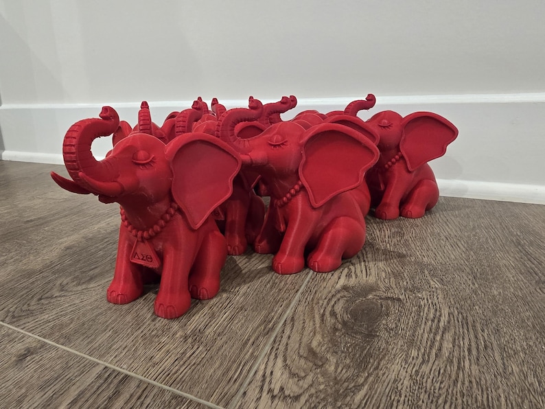 May include: A collection of red elephant figurines. Each figurine has large ears, a curled trunk, and a beaded necklace. The elephants are sitting on a wooden surface. The figurines have closed eyes.