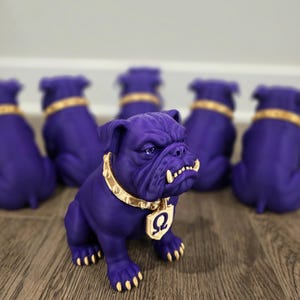 May include: A purple bulldog figurine with a matte finish, featuring a metallic gold spiked collar and a gold pendant with a Greek Omega symbol. The bulldog has gold-painted claws and teeth, and is shown in a group of identical purple figurines.