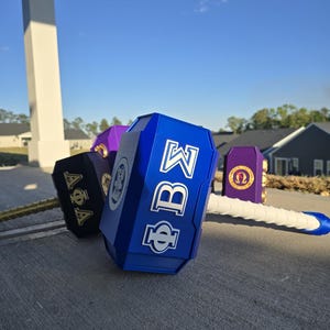 May include: A collection of decorative hammers in various colors, including blue, black, and purple. Each hammer features Greek letters and symbols, with a white and gold handle. The hammers are displayed on a gray surface against a blue sky.