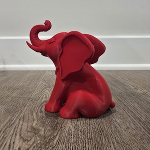 May include: A red elephant figurine with its trunk raised and eyes closed. The elephant is sitting on its hind legs. Another red elephant figurine is partially visible in the background.