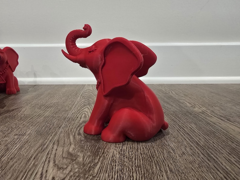 May include: A red elephant figurine with its trunk raised and eyes closed. The elephant is sitting on its hind legs. Another red elephant figurine is partially visible in the background.