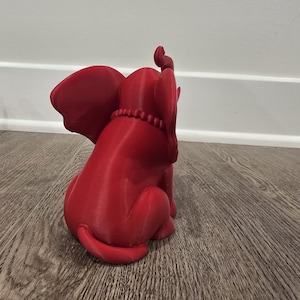 May include: A red elephant figurine with large ears and a beaded necklace. The elephant is in a sitting position, with its trunk raised. Another similar figurine is partially visible in the background. The figurine is made of a solid material.