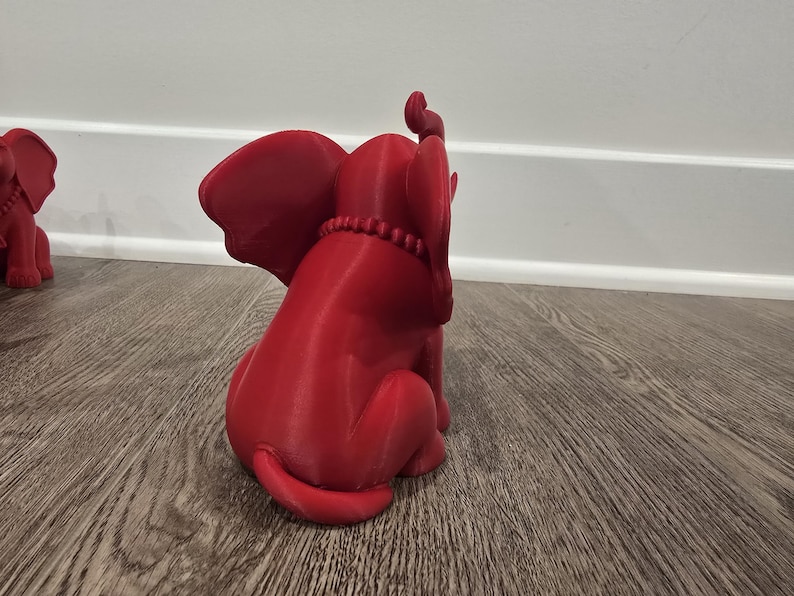May include: A red elephant figurine with large ears and a beaded necklace. The elephant is in a sitting position, with its trunk raised. Another similar figurine is partially visible in the background. The figurine is made of a solid material.