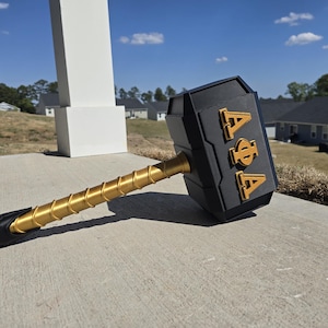 May include: A black and gold decorative hammer with the Greek letters A, Φ, and A. The hammer head is black with gold lettering. The handle is gold with a black grip. The hammer is resting on a concrete surface.