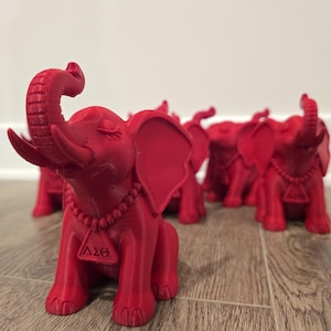 May include: A collection of red elephant figurines. Each elephant has its trunk raised and wears a beaded necklace with a triangular pendant. The figurines are arranged on a wooden surface.