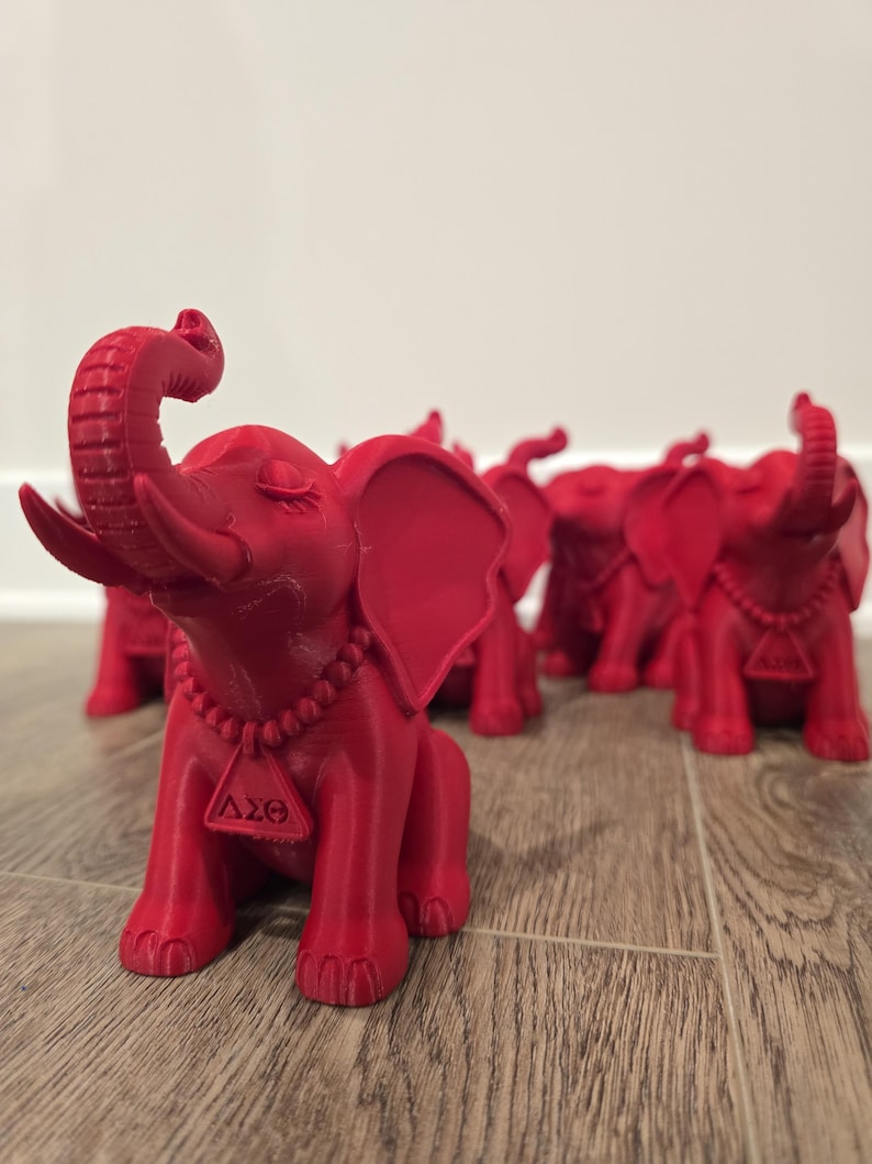 May include: A collection of red elephant figurines. Each elephant has its trunk raised and wears a beaded necklace with a triangular pendant. The figurines are arranged on a wooden surface.