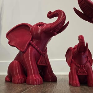 May include: A set of three red 3D-printed elephant figurines in varying sizes. Each elephant features a raised trunk, large ears, and a beaded necklace with a triangular pendant displaying the Greek letters Delta Sigma Theta, or &Delta;&Sigma;&Theta;.