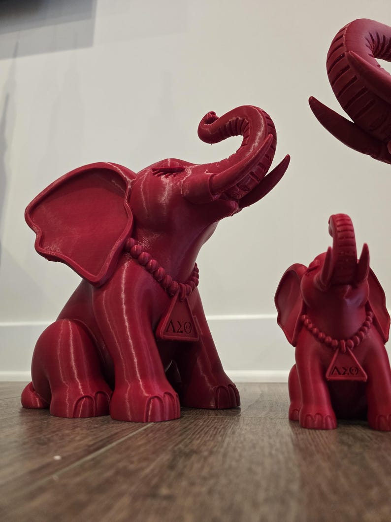 May include: A set of three red 3D-printed elephant figurines in varying sizes. Each elephant features a raised trunk, large ears, and a beaded necklace with a triangular pendant displaying the Greek letters Delta Sigma Theta, or &Delta;&Sigma;&Theta;.