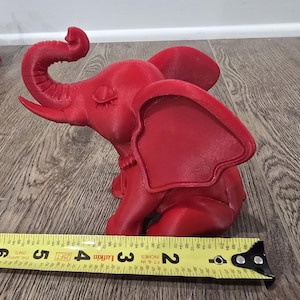 May include: A red elephant figurine, approximately 5 inches long, sits on a wooden surface. The elephant has large ears, a curled trunk, and is in a seated position. A yellow measuring tape is in the foreground.
