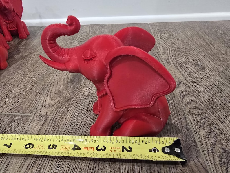 May include: A red elephant figurine, approximately 5 inches long, sits on a wooden surface. The elephant has large ears, a curled trunk, and is in a seated position. A yellow measuring tape is in the foreground.