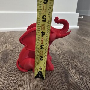 May include: A red elephant figurine, approximately 5 inches tall, is measured with a yellow tape measure. The figurine is sitting on a wooden floor. The elephant has large ears and a curled trunk.