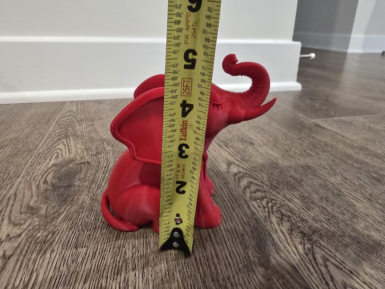 May include: A red elephant figurine, approximately 5 inches tall, is measured with a yellow tape measure. The figurine is sitting on a wooden floor. The elephant has large ears and a curled trunk.
