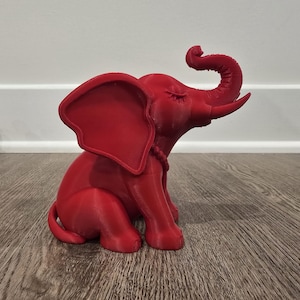 May include: A red elephant figurine with its trunk raised and eyes closed. The elephant is sitting on its hind legs, with large ears and a rope-like detail around its neck. The figurine is on a wooden surface.