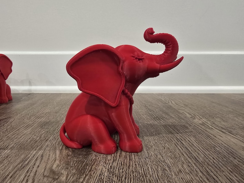 May include: A red elephant figurine with its trunk raised and eyes closed. The elephant is sitting on its hind legs, with large ears and a rope-like detail around its neck. The figurine is on a wooden surface.