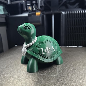 May include: A 3D-printed green turtle figurine featuring a white beaded necklace with a flower pendant. The turtle shell displays the Greek letters Iota, Phi, and Lambda in white. The surface of the turtle shows visible horizontal print layers.